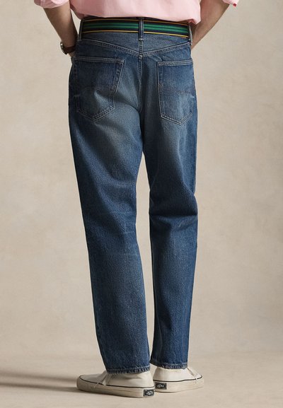 Άτομο που φοράει μπλε τζιν, λευκά αθλητικά παπούτσια και ροζ πουκάμισο, στέκεται μπροστά σε μπεζ φόντο, ορατό από πίσω.