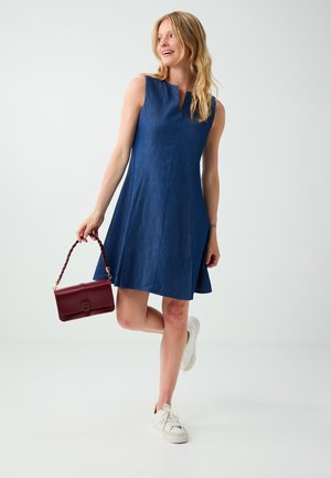 Femme portant une robe bleue sans manches et des baskets blanches, tenant un petit sac à main rouge, debout sur une jambe devant un fond uni.