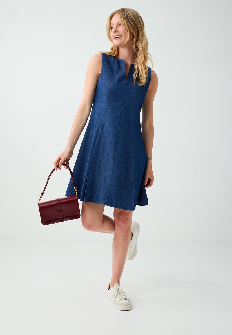 Femme portant une robe bleue sans manches et des baskets blanches, tenant un petit sac à main rouge, debout sur une jambe devant un fond uni.