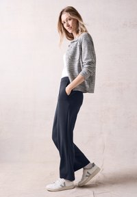 Grå texturerad kofta, vit linne, marinblå vida byxor och vita sneakers med grå accenter. Modell poserar med händerna i fickorna.