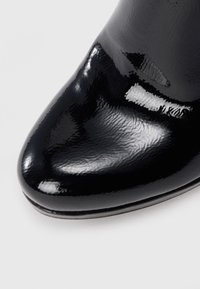 Bout de chaussure en cuir verni noir brillant avec surface lisse et réfléchissante et embout arrondi sur un fond clair.