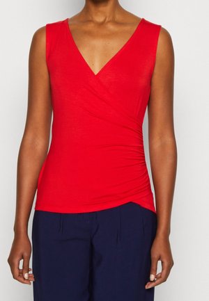 Vrouw draagt een mouwloze rode wikkeltop met plooien aan één kant en marineblauwe broek, staand tegen een effen achtergrond.