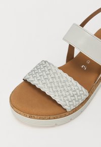 White sandals with a woven silver strap, a tan cork footbed, and a white rubber sole. The strap has a scalloped edge detail.