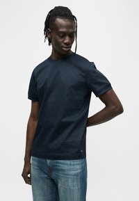 Μπλε ναυτικό κοντομάνικο βαμβακερό t-shirt με στρογγυλή λαιμόκοψη, λεία υφή και μικρή λεπτομέρεια με λογότυπο κοντά στη βάση. Φορεμένο με ανοιχτόχρωμα τζιν.