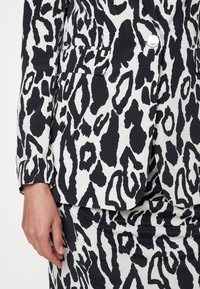 Blazer et jupe assortis à imprimé animal abstrait noir et blanc, avec un bouton argenté et une poche latérale, portés par une personne avec une main visible.