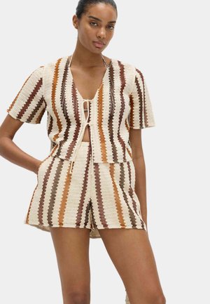 Kobieta ubrana w beżowy, brązowy i pomarańczowy pasiasty dzianinowy top z krótkim rękawem oraz pasujące, wysokie szorty, stojąca z jedną ręką na biodrze.