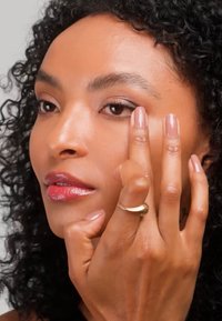 Donna con capelli ricci applica il trucco sulla palpebra usando due dita, mostrando pelle liscia, unghie naturali e un anello d'oro al dito.