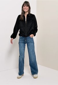 Czarna, miękka kurtka na zamek z dwoma kieszeniami na piersi i elastycznymi mankietami, zestawiona z jasnoniebieskimi jeansami o szerokich nogawkach i beżowymi butami.