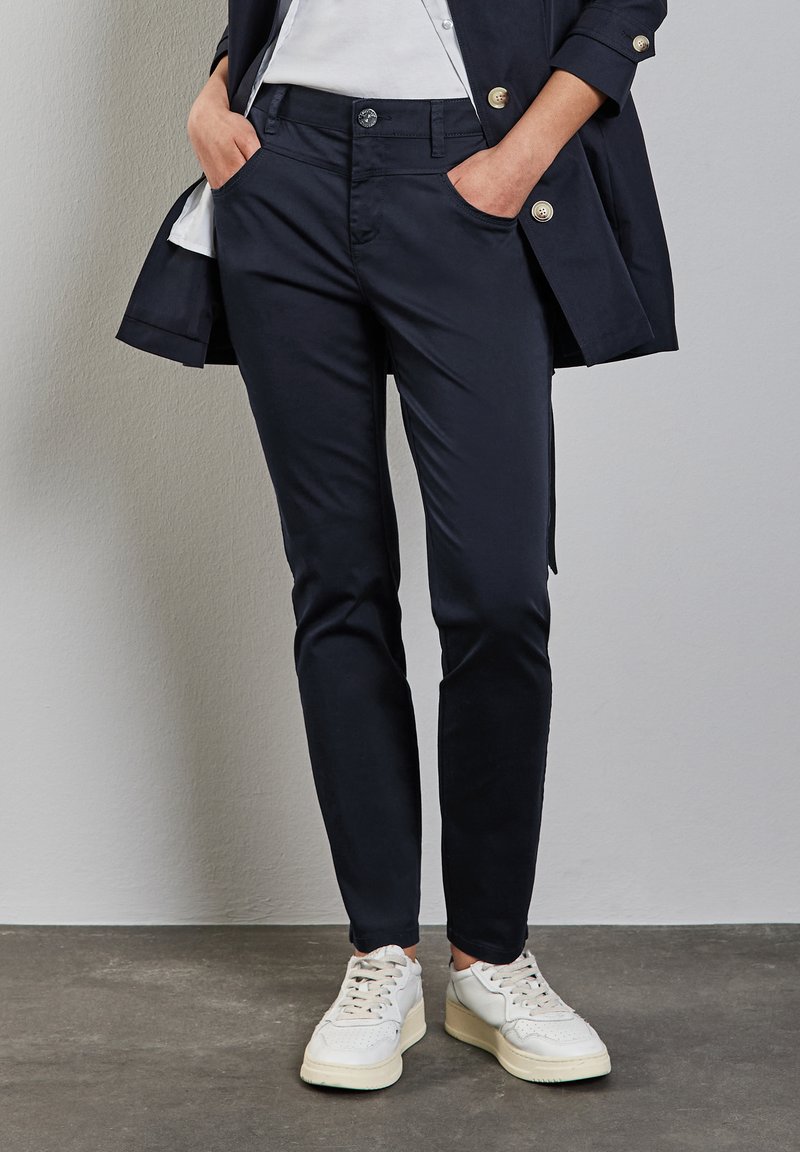 Pantalones ajustados de color azul marino, hechos de una mezcla de algodón, con bolsillos delanteros y cierre de botón. Combinados con zapatillas blancas.
