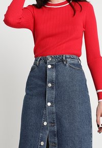 Haut rouge à côtes avec des manches longues et un col rond blanc, associé à une jupe en denim bleu arrivant au genou avec une fermeture à boutons sur le devant et des poches.