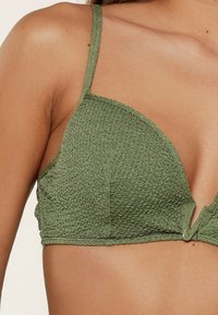 Bralette verde, con texture, dotato di spalline sottili, coppe triangolari e dettaglio con apertura frontale. Il materiale presenta una leggera lucentezza.