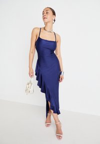 Navy blue satin dress with spaghetti straps, asymmetric ruffle detail, thigh-high slit, and smooth texture. Model holds a small beige bag.