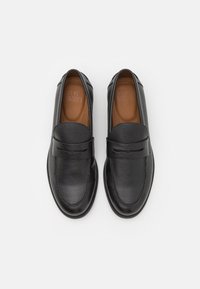 Mocassins en cuir noir à finition texturée, avec un détail de large bande, un bout en forme d'amande et une semelle intérieure lisse marron. Semelle en caoutchouc profil bas.
