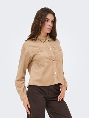 Kvinde iført en beige skjorte med knapper og brystlommer samt mørke bukser, poserer med den ene hånd i lommen og kigger ud til siden.