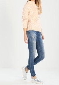 Ljusrosa kabelstickad tröja med hög krage, i kombination med blå slitna slim-fit jeans och silverfärgade slip-on sneakers.