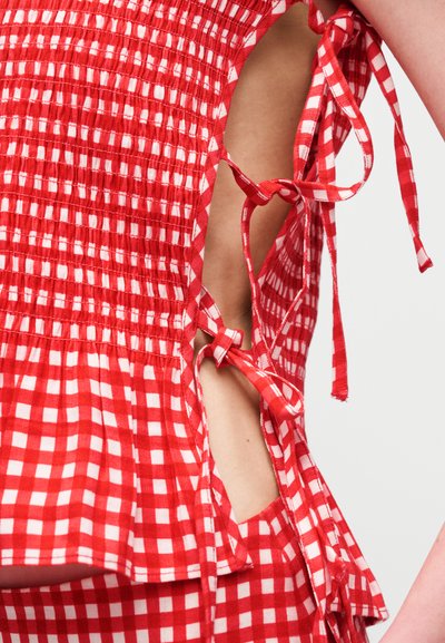 Ενδυμα με κόκκινο και λευκό καρό σχέδιο, με πλαϊνές ανοίγματα δεμένα με υφασμάτινα φιόγκους, που αποκαλύπτουν δέρμα στο πλευρικό τμήμα του κορμού ενός ατόμου.