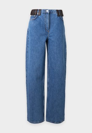 Kvinde med kort mørkt hår iført en sort langærmet top, vidde blå jeans med sort bælte og sorte sko, stående mod en ensfarvet baggrund.