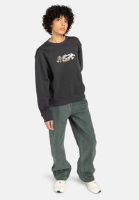 Zwarte sweatshirt met kleurrijke grafische borduursels, ontspannen pasvorm, geribbelde manchetten en zoom, gecombineerd met losse groene jeans en witte sneakers.