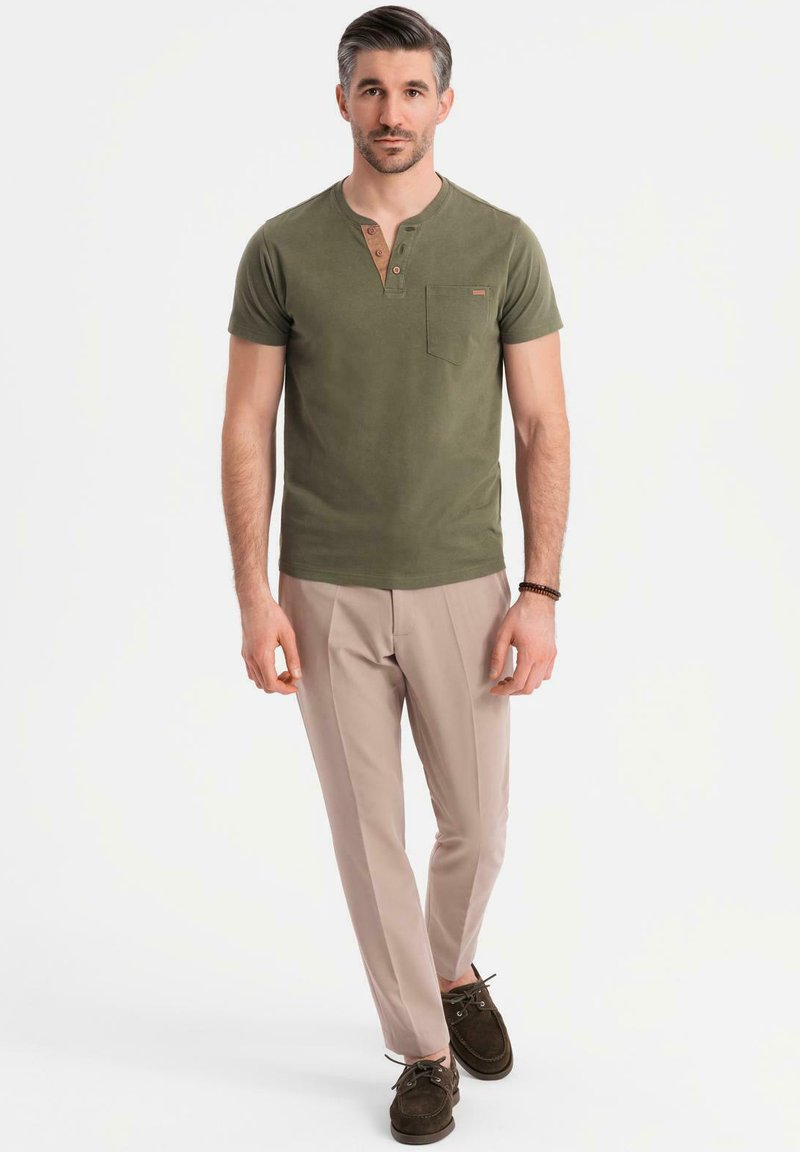 Oliwkowa, krótko rękawowa koszulka typu henley z kieszonką na piersi i drewnianymi guzikami, w połączeniu z jasnobeżowymi spodniami i ciemnymi butami.