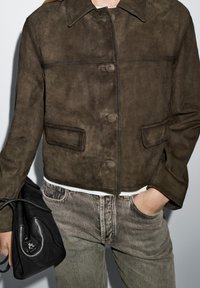 Brown suede jacket with a cropped design, button closures, and two front pockets. Paired with gray jeans and a black handbag.