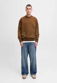 Ung mand stående iført brun sweatshirt, hvid undertrøje, brede blå jeans og beige mokkasinstil-sko på hvid baggrund.