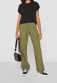 Volné olivově zelené cargo kalhoty s bočními kapsami, kombinované s černým topem s krátkým rukávem a černými teniskami, držící malou černou tašku.