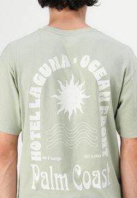 Lys grøn bomulds t-shirt med et stort hvidt solgrafik, tekst der læser "Hotel Laguna Ocean Front," og bølgede linjer design på ryggen.