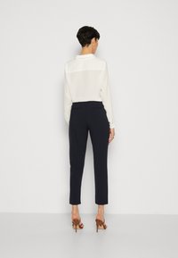 Bela bluza z dolgimi rokavi s ovratnikom, kombinirana z mornarsko modrimi krojenimi hlačami in rjavimi čevlji s peto. Gladek material, minimalni detajli.