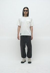 Mann med skulderlangt hår iført svarte solbriller, hvit t-skjorte med lommer, svarte bukser og beige sneakers som står mot en helt hvit bakgrunn.