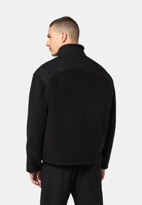 Schwarze Fleecejacke mit hohem Kragen, kontrastierendem Frontpaneel, bequemer Passform und sichtbarer Textur, kombiniert mit schwarzen Hosen.
