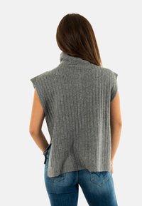 Top de punto sin mangas de canalé gris con cuello alto, que presenta un corte relajado y slit laterales, combinado con vaqueros.