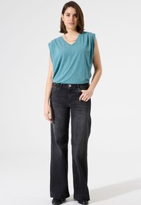 Teal V-Ausschnitt ärmelloses Oberteil, kombiniert mit hoch taillierten schwarzen Schlagjeans, die einen Knopfverschluss und eine dezente Rippstruktur aufweisen.