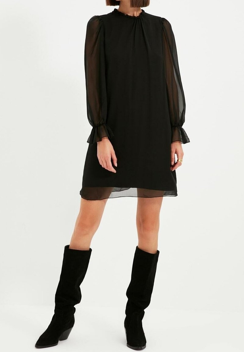 Schwarzes Mini-Kleid mit langen, durchsichtigen Ärmeln, gerafftem Ausschnitt und Rüschenmanschetten. Getragen mit knielangen Stiefeln aus schwarzem Wildleder. Glatte Textur.