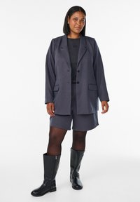 Tmavošedý blazer s dvomi gombíkmi a kapsami s chlopňami, v kombinácii s rovnako sfarbenými šortkami. Doplnené čiernymi vysokými čižmami po kolená. Hladká textúra tkaniny.