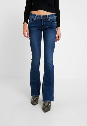 Kobieta w ciemnoniebieskich jeansach bootcut, czarnej bluzce z długim rękawem oraz szpiczastych botkach z wzorem skóry węża, stojąca na jednolitym białym tle.