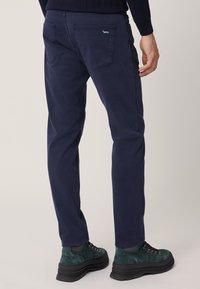Pantaloni in cotone blu navy con vestibilità slim, dotati di due tasche posteriori e un piccolo logo a forma di cane blu. Indossati con stivali da escursionismo verdi.