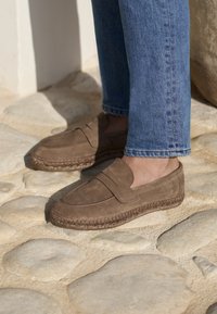 Scarpe slip-on in camoscio marrone con suola intrecciata in espadrillas, caratterizzate da una texture liscia e accenti decorativi minimi, posizionate su ciottoli.