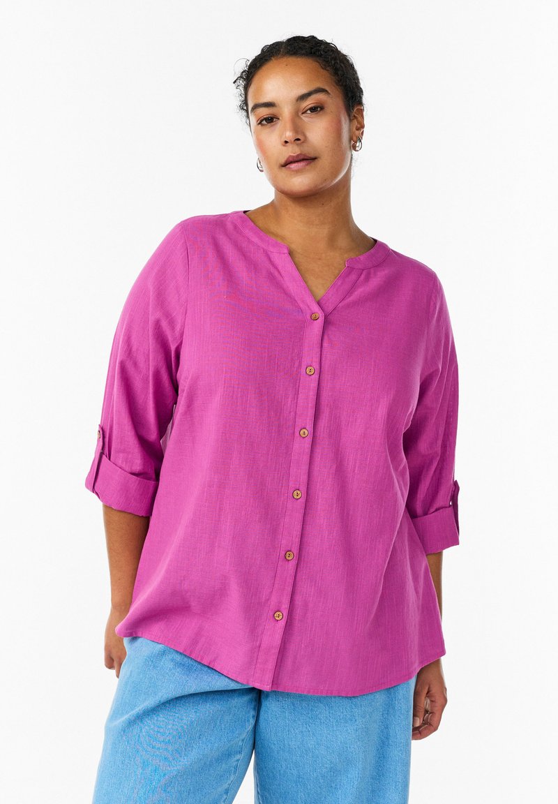 Fialová košeľa na gombíky s výstrihom do V, rukávmi na vyhrnutie a drevenými gombíkmi. Textúrovaná látka, voľný strih, kombinovaná so svetlomodrými nohavicami.
