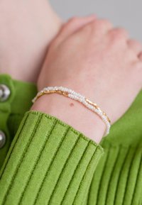 Bracelet en perles dorées et blanches sur un poignet, avec de délicates accents dorés et de petites perles blanches, sur fond d'un pull en maille verte.