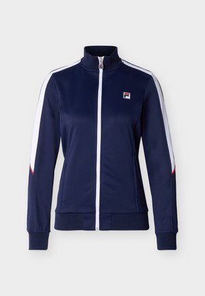 Veste zippée bleu marine avec des rayures blanches sur les manches, des accents rouges près des poignets, un ourlet et des poignets côtelés, et un petit logo sur le côté gauche de la poitrine.