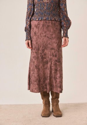 Mujer con una falda larga texturizada en color malva, blusa de manga larga con estampado floral, cintura y mangas fruncidas, y botas de ante marrones hasta media pantorrilla.