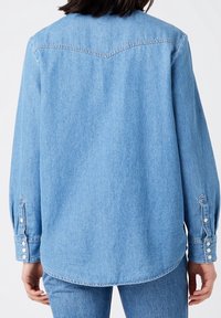 Light blue denim shirt with long sleeves, slightly curved hem, and snap-button cuffs. Features a back yoke seam design.