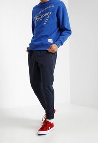 Felpa blu con logo bianco, pantaloni neri e sneakers rosse con accenti bianchi. Il materiale è probabilmente cotone; design casual con vestibilità rilassata.