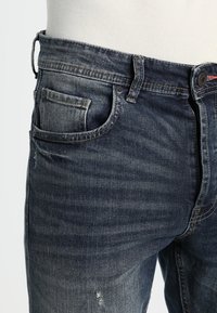 Tummansiniset farkut, joissa on klassinen lahje, etutasku ja kulutusta korostava yksityiskohta, teksturoitu kangas, jossa on hienosti haalennutta ja kulumaa.