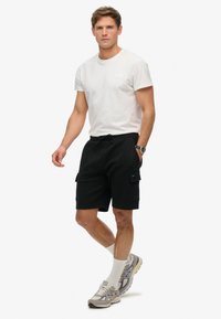 Czarne szorty cargo z bocznymi kieszeniami, w połączeniu z gładką białą koszulką. Model nosi szare i białe sneakersy z wysokimi skarpetkami.