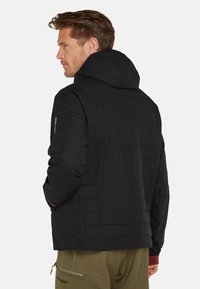 Mężczyzna z krótkimi brązowymi włosami, ubrany w czarną pikowaną kurtkę z kapturem i oliwkowe spodnie, stojący bokiem na białym tle.