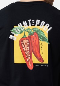 Sort t-shirt med et dristigt grafisk design af røde chili peberfrugter på en gul baggrund, med teksten "BRIGHT=POOL" ovenfor og "Just chilling" nedenfor.