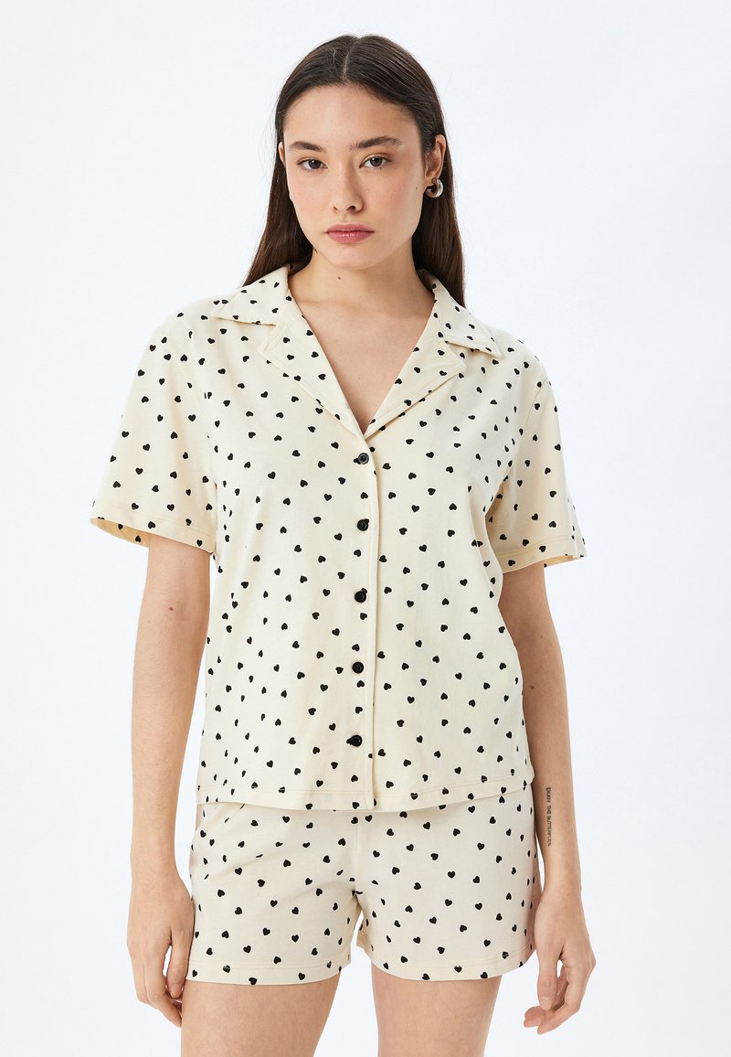 Kortärmad beige skjorta och shorts-set med svart hjärttryck, som har krage och knapplist framtill för en avslappnad look.