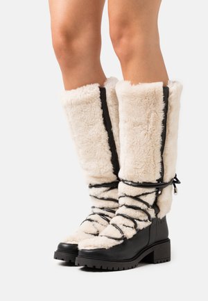 Høje sorte støvler med tyk beige lammeskindsfôr, snoet med sorte snørebånd, båret på bare ben mod en ensfarvet lys baggrund.