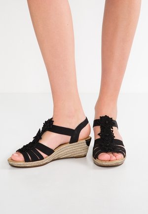 Pieds portant des sandales compensées noires avec des semelles tressées et des décorations florales sur les brides, sur fond blanc.